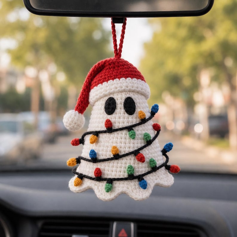 May include: A white crocheted ghost ornament wearing a red Santa hat with a white pom-pom. The ghost is wrapped in a black string of miniature multi-colored Christmas lights in red, green, blue, and yellow, hanging from a red crocheted loop.