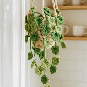 May include: A crocheted hanging plant holder with trailing green leaves. The beige pot is suspended by beige rope. The leaves are various shades of green, creating a natural, handmade aesthetic. The plant holder is designed to hold a small plant.