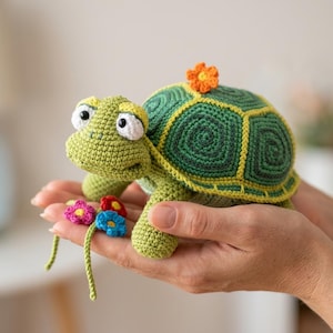Könnte beinhalten: Ein kleines, handgefertigtes Häkel-Schildkrötenspielzeug in hellen und dunklen Grüntönen. Die Schildkröte hat einen strukturierten Panzer mit einer orangefarbenen Blume oben, große weiße Augen mit schwarzen Pupillen und einen kleinen Strauß aus rosa, roten und blauen Blumen.