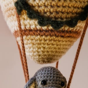 May include: A handmade crochet hot air balloon with a gray elephant in the basket. The balloon is beige, brown, and green, with brown rope suspending the basket. The elephant has gray fur and black eyes. A whimsical, handcrafted toy.