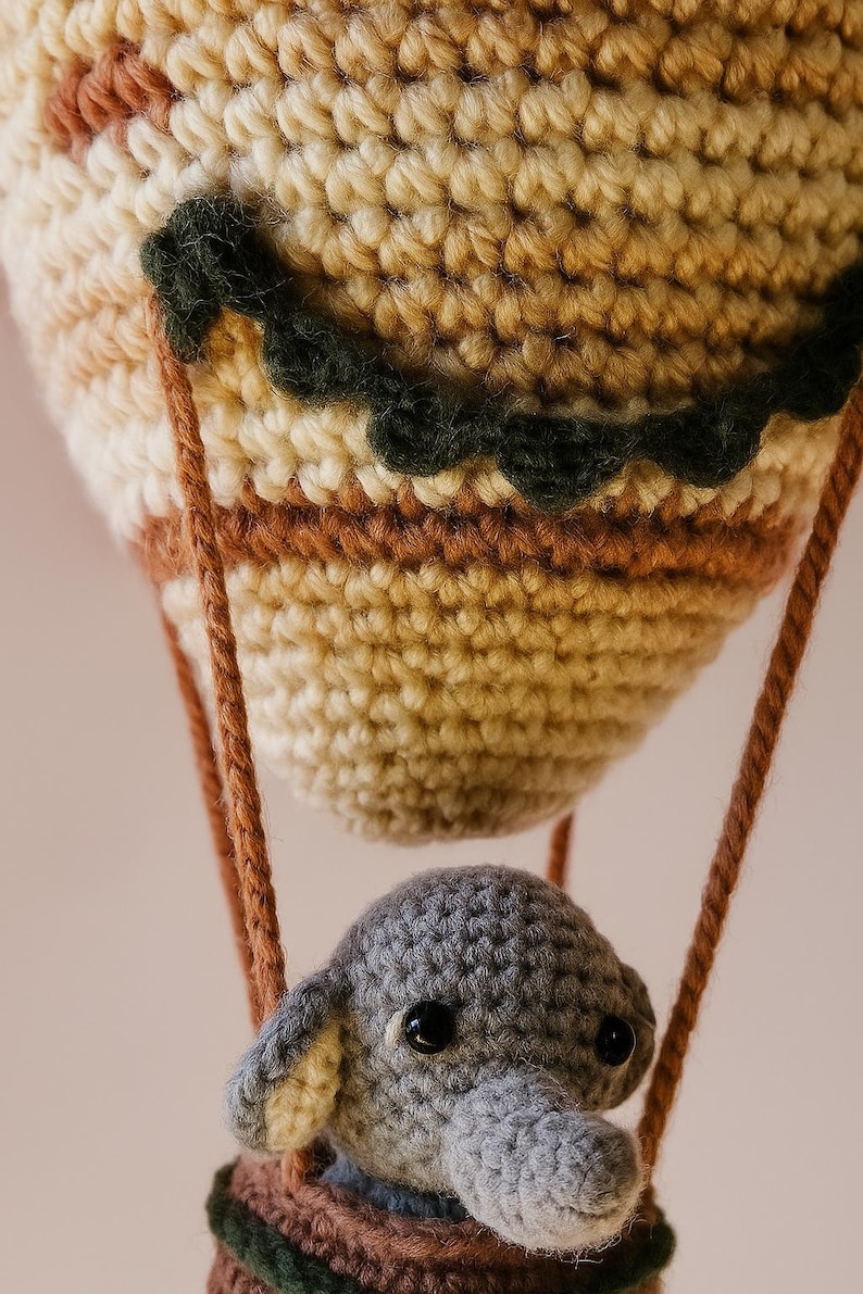 May include: A handmade crochet hot air balloon with a gray elephant in the basket. The balloon is beige, brown, and green, with brown rope suspending the basket. The elephant has gray fur and black eyes. A whimsical, handcrafted toy.