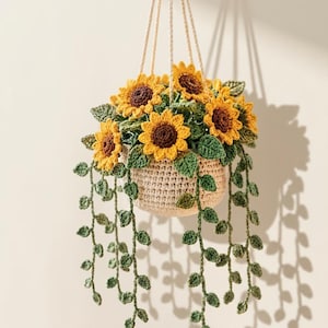 May include: A crocheted hanging plant featuring bright yellow sunflowers with dark brown centers, green leaves, and long trailing vines. The plant is contained in a beige crocheted basket suspended by cream-colored cords, creating a decorative display.