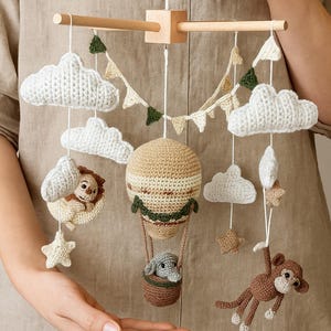 May include: A handmade crochet baby mobile featuring a hot air balloon, clouds, stars, and animal figures. The mobile includes a lion, elephant, and monkey. The color palette consists of beige, brown, white, and green.