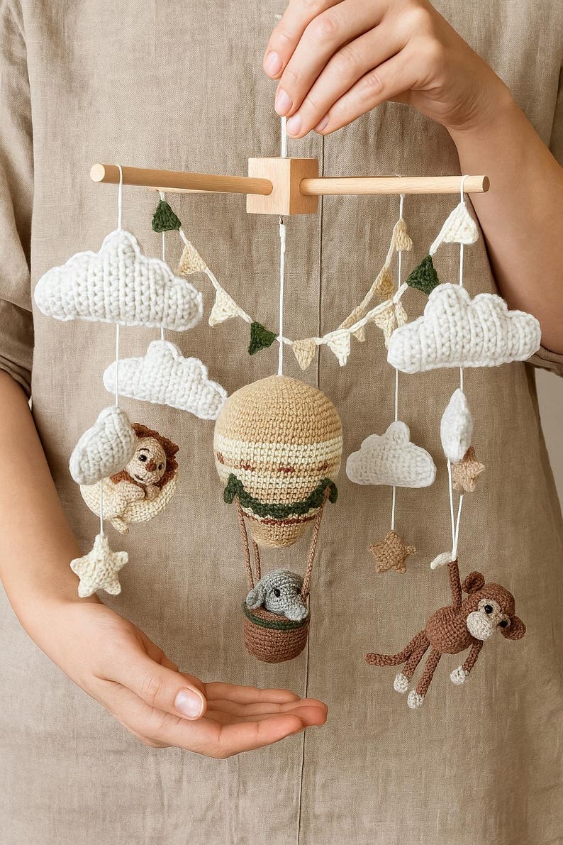 May include: A handmade crochet baby mobile featuring a hot air balloon, clouds, stars, and animal figures. The mobile includes a lion, elephant, and monkey. The color palette consists of beige, brown, white, and green.
