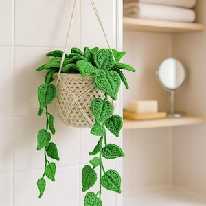 May include: A handmade crochet hanging planter with vibrant green leaves and a beige pot. The plant is suspended by a cream-colored rope, adding a touch of nature to any space. The leaves cascade down, creating a charming, decorative accent.