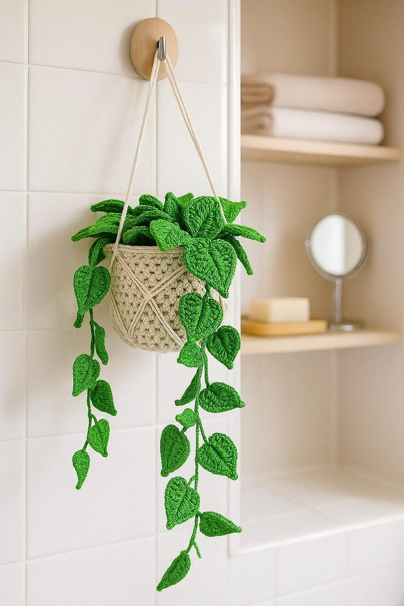 May include: A handmade crochet hanging planter with vibrant green leaves and a beige pot. The plant is suspended by a cream-colored rope, adding a touch of nature to any space. The leaves cascade down, creating a charming, decorative accent.