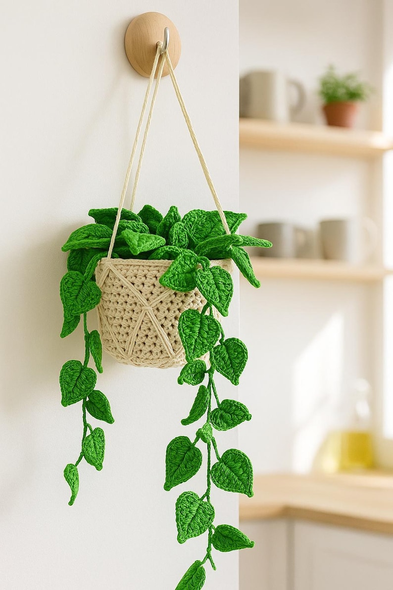 May include: A handmade crochet hanging planter with vibrant green leaves and a beige woven basket. The plant is suspended from a wooden wall hook with cream-colored cords, adding a touch of nature to interior decor.