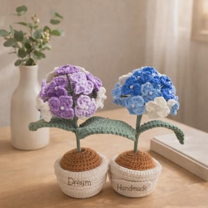 Puede incluir: Dos flores de hortensia hechas a mano con ganchillo en macetas pequeñas. Una flor es morada y blanca, la otra azul y blanca. Las macetas son beige con las palabras "Dream" y "Handmade". Las flores están sobre una mesa de madera.