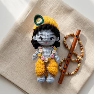 Könnte beinhalten: Handgefertigte Krishna-Häkelpuppe mit blauer Haut, gelber Kleidung und einem dekorativen Kopfschmuck. Eine Holzflöte und eine Perlenkette liegen daneben. Die Puppe ist etwa 15 cm groß.