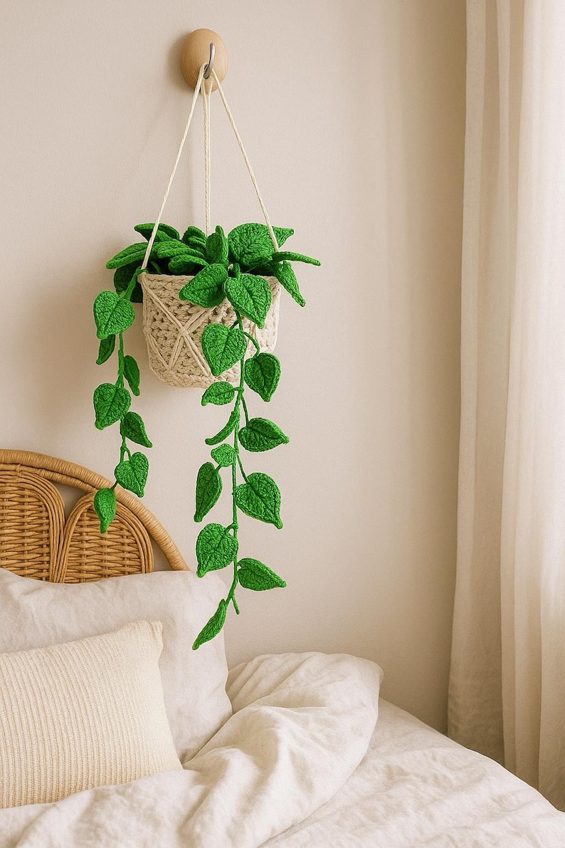 May include: A handmade crochet hanging plant with vibrant green leaves and a beige woven basket. The plant is suspended from a wooden hook on a light-colored wall, adding a touch of nature to the room. The leaves cascade downwards, creating a decorative accent.