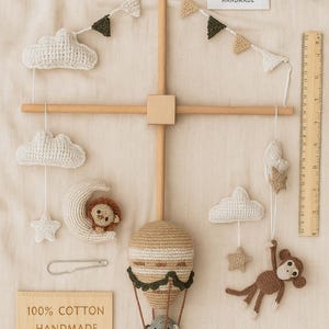 May include: A handmade baby mobile featuring crocheted clouds, stars, a moon with a lion, a hot air balloon with a koala, and a monkey. The mobile is made of beige, brown, and white yarn, with wooden accents. The text "100% Cotton Handmade" is visible.