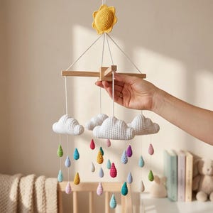 May include: A handmade crochet baby mobile featuring a yellow sun, white clouds, and colorful raindrops. The mobile is suspended from a wooden frame and is designed for a nursery or baby's room.
