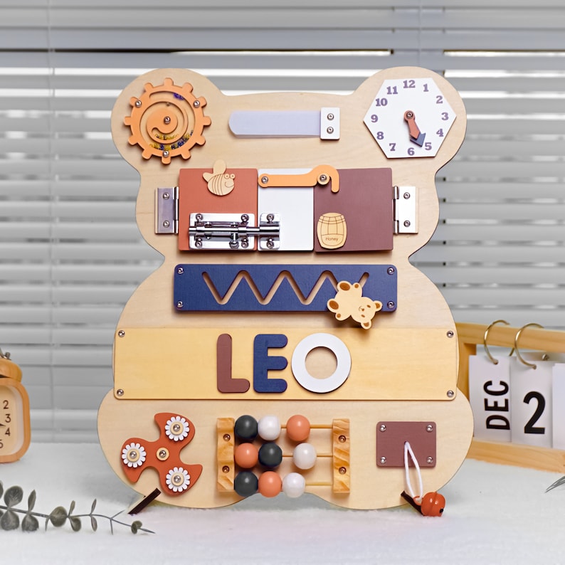 May include: Wooden bear-shaped busy board with various activities for toddlers, including a clock, a sliding latch, a shape sorter, a bead maze, a spinning gear, and a name puzzle that reads "LEO".