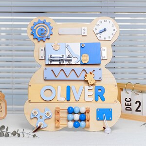May include: Wooden activity board shaped like a bear with various textures and activities for children. The board features a clock, a sliding door, a latch, a spinning gear, a bead maze, a shape sorter, and a spinning fidget toy. The board is personalized with the name "OLIVER".