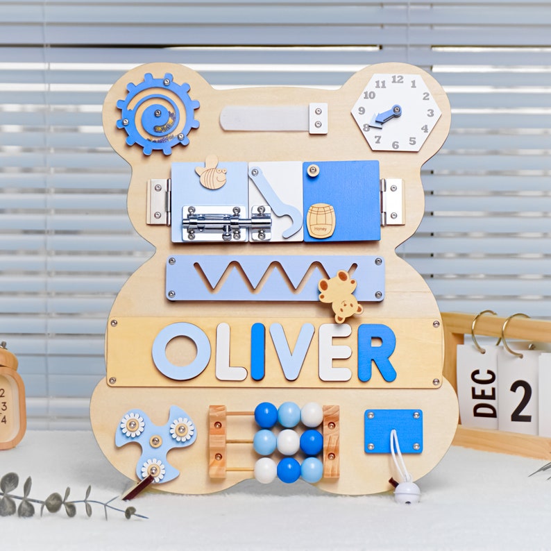 May include: Wooden activity board shaped like a bear with various textures and activities for children. The board features a clock, a sliding door, a latch, a spinning gear, a bead maze, a shape sorter, and a spinning fidget toy. The board is personalized with the name "OLIVER".