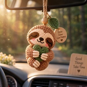 May include: A crocheted sloth car ornament in shades of brown and cream, holding a small green heart. It features a wooden bead, a green crocheted leaf, and a round wooden tag engraved with the text Drive Slow Live Happy and a heart symbol.