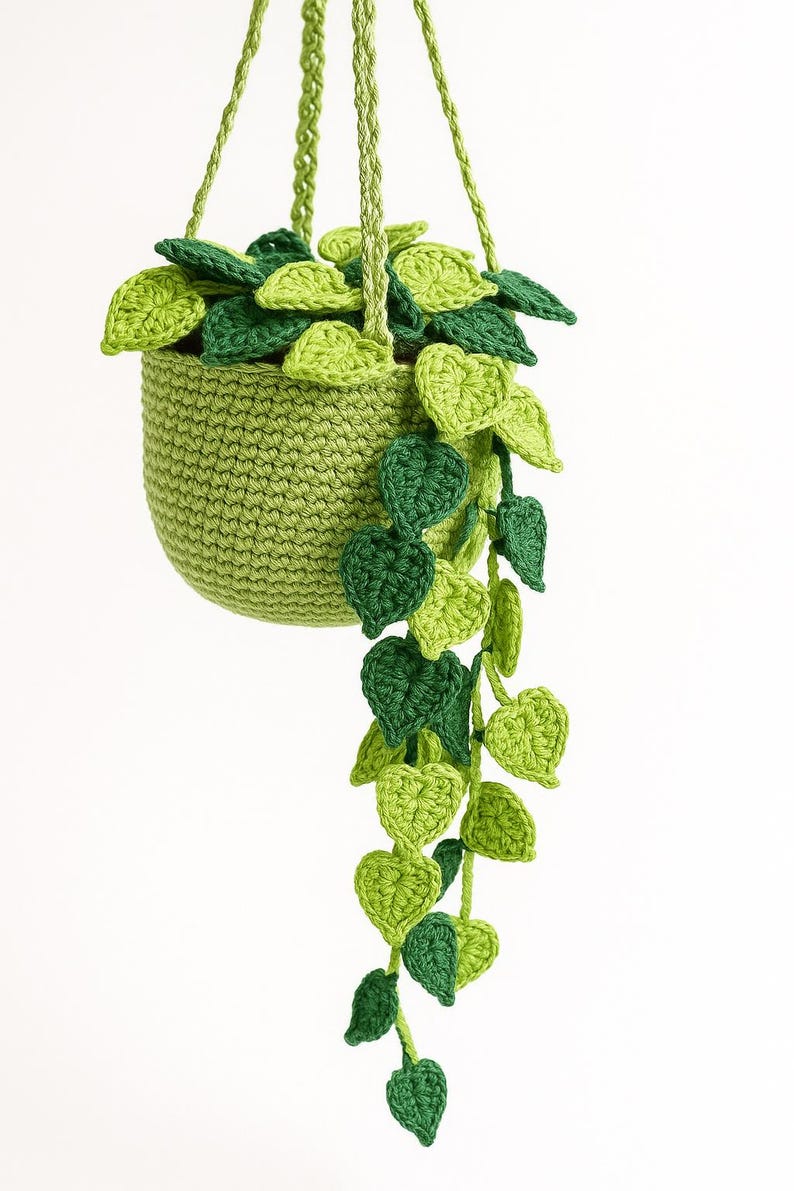 May include: A handmade crochet hanging planter with a trailing plant. The planter is a solid light green, with a woven texture. The plant has various shades of green leaves, creating a natural, textured look. The hanging cords are also light green.