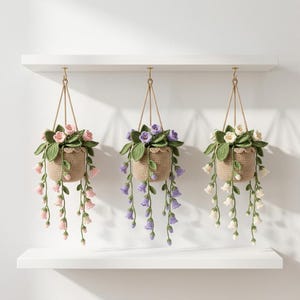 Può includere: Tre piante pensili all'uncinetto appese a una mensola. Ogni cestino color marrone chiaro contiene foglie verdi e fiori a forma di campanula che ricadono in tonalità rosa chiaro, viola e bianco, realizzati con trame di filato dettagliate.