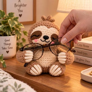 May include: A crocheted sloth figurine in shades of light and dark brown with cream-coloured accents on the face, belly, and paws. The sloth has a small tuft of hair on its head, closed eyes, and a black nose, designed to hold a pair of tortoiseshell glasses.