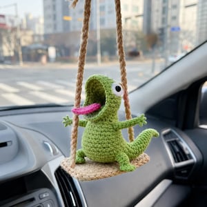 Könnte beinhalten: Eine grüne, gehäkelte Echsenfigur mit weit geöffnetem Maul, herausgestreckter rosa Zunge und großen weißen Augen. Die Echse sitzt auf einer kleinen, beigen, gehäkelten Plattform, die an zwei dicken, geflochtenen beigen Seilen aufgehängt ist.