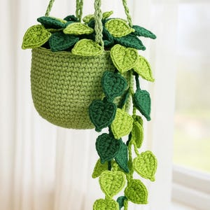 May include: A handmade crochet hanging planter in shades of green. The planter is filled with crochet leaves in varying shades of green, with trailing vines of leaves cascading down. The planter is suspended by a green rope.