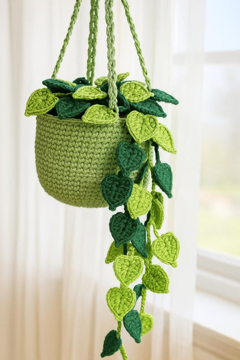 May include: A handmade crochet hanging planter in shades of green. The planter is filled with crochet leaves in varying shades of green, with trailing vines of leaves cascading down. The planter is suspended by a green rope.