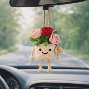 May include: A crocheted car charm shaped like a smiling flower pot with a red and pink rose arrangement. The beige pot has arms and legs, and a wooden tag that says "Be safe". The charm hangs from the rearview mirror.