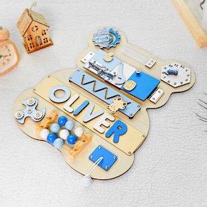 May include: A wooden activity board with a blue and beige color scheme. The board features a variety of activities for children, including a clock, a bead maze, a spinning fidget toy, a sliding latch, a shape sorter, and a name puzzle that reads "OLIVER".