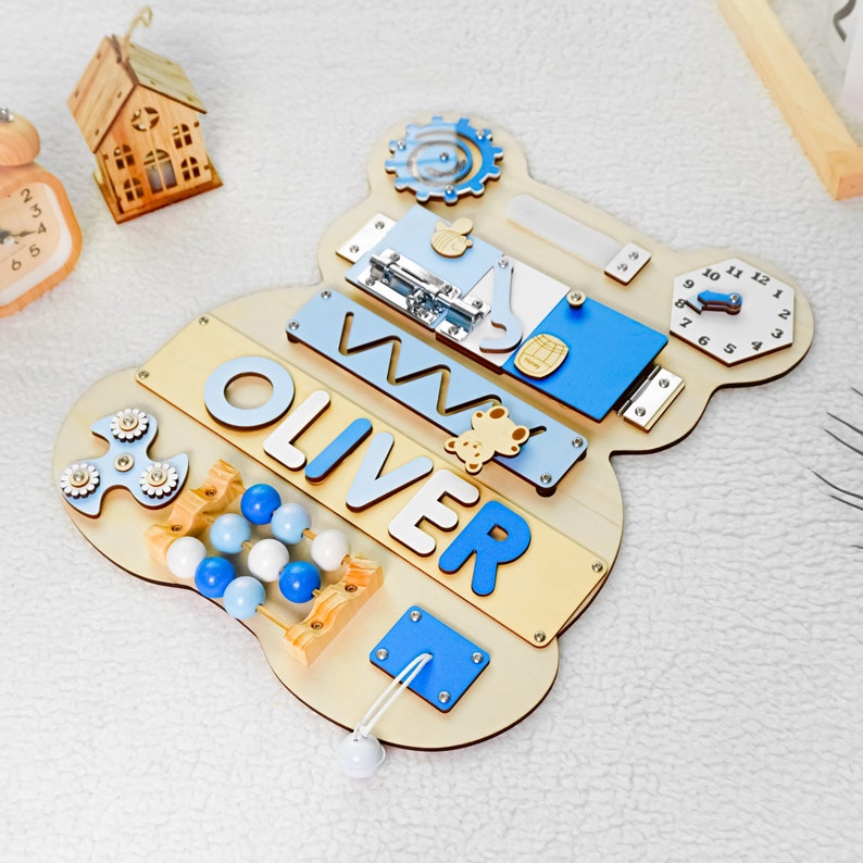 May include: A wooden activity board with a blue and beige color scheme. The board features a variety of activities for children, including a clock, a bead maze, a spinning fidget toy, a sliding latch, a shape sorter, and a name puzzle that reads "OLIVER".