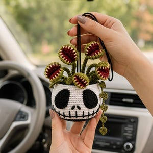 May include: A crocheted hanging ornament featuring a white pot with a black skull face design, topped with several green and dark red Venus flytrap plants. A long, thin green vine with small heart-shaped leaves hangs down from the side of the pot.