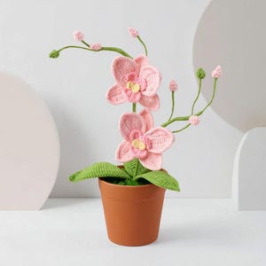 Op de afbeelding: Een gehaakte roze orchidee in een bruine pot. De handgemaakte bloem heeft gedetailleerde bloemblaadjes met gele centra, groene bladeren en stelen. De plant is een decoratief item, perfect om een vleugje natuur toe te voegen.
