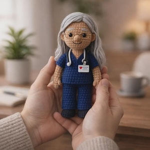 May include: A crocheted doll of a healthcare worker, wearing a blue uniform, glasses, and a stethoscope. The doll has gray hair and a name tag with a heart symbol. The doll is held in two hands.
