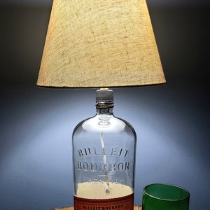 May include: A clear glass bottle lamp with a brown fabric lampshade. The bottle is labeled "BULLEIT BOURBON FRONTIER WHISKEY". The lamp is sitting on a wooden surface next to a green glass.