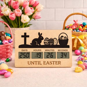 May include: Wooden Easter countdown timer with a black silhouette of a cross, bunny, and Easter eggs. The digital display shows 250 days, 19 hours, 26 minutes, and 34 seconds until Easter. Baskets of Easter eggs and flowers are in the background.