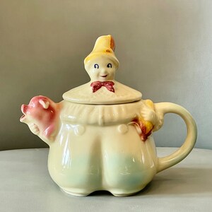 Puede incluir: Una tetera de cerámica vintage con forma de persona que lleva un sombrero amarillo y sostiene un cerdo rosa. La tetera tiene un diseño caprichoso y está pintada en tonos de amarillo, rosa y azul.