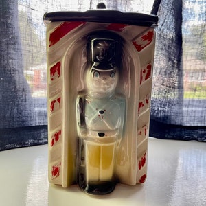 May include: A vintage ceramic cookie jar shaped like a toy soldier standing in a doorway. The soldier is painted in shades of blue, yellow, and black. The doorway is painted in red and white stripes.