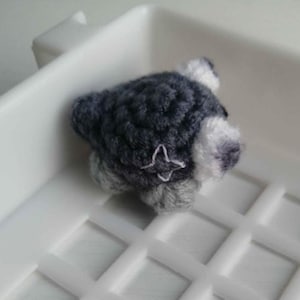 Puede incluir: Una figura de perro en miniatura hecha a mano, tejida a ganchillo, en tonos grises y blancos. El perro tiene un detalle en forma de estrella en un costado. La figura est&aacute; sobre una superficie blanca.