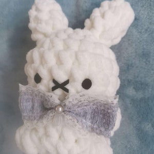 May include: White crocheted bunny with a gray and white bow and a pearl accent. The bunny has black eyes and a black X for a mouth.