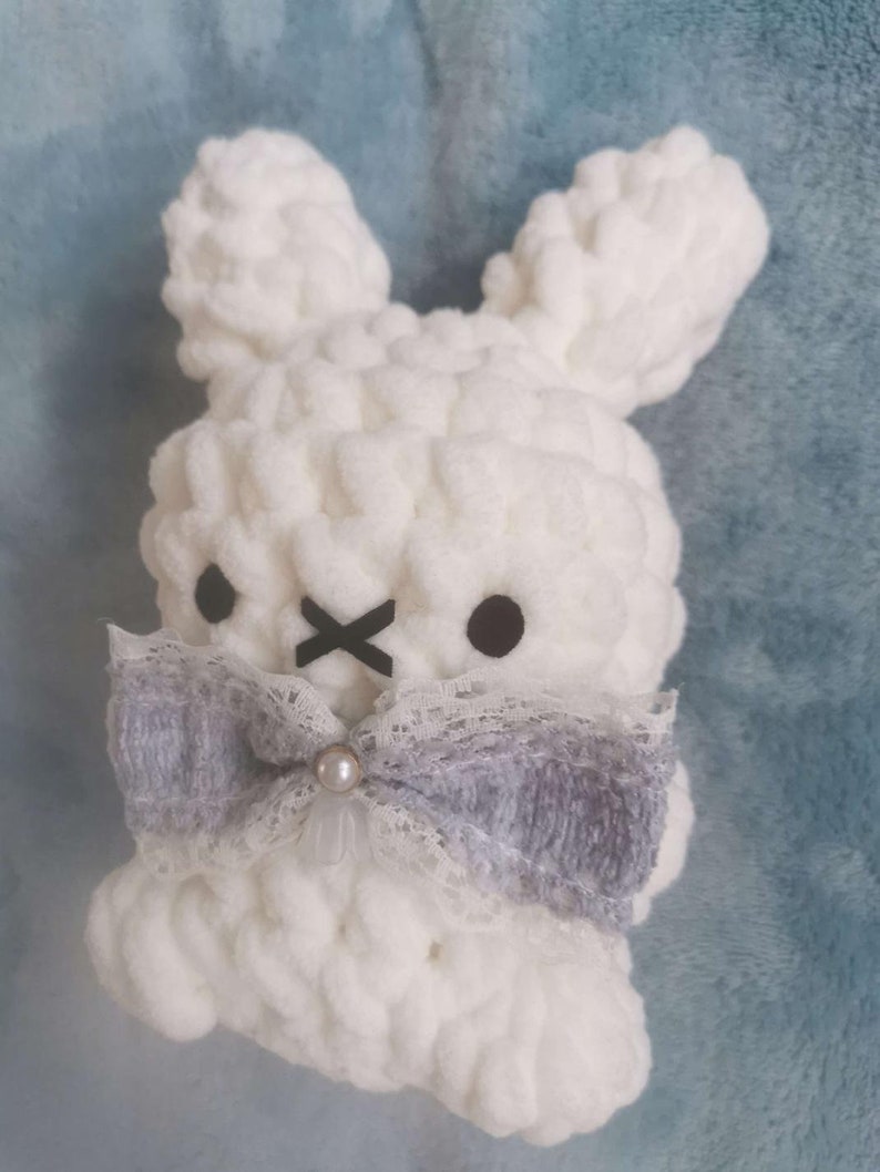 May include: White crocheted bunny with a gray and white bow and a pearl accent. The bunny has black eyes and a black X for a mouth.