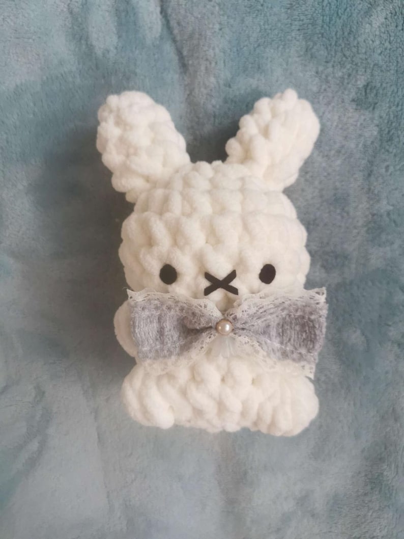 May include: White crocheted bunny with gray bow and pearl accent. The bunny has black eyes and a stitched X for a mouth.