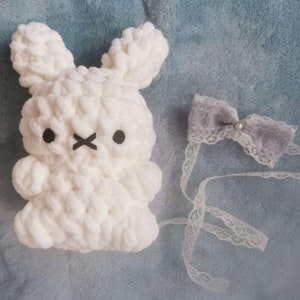 May include: White crocheted bunny plush toy with black eyes and a stitched X-shaped mouth. The bunny is wearing a gray bow with a white pearl in the center, attached to a white lace ribbon.