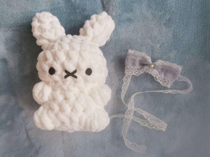 May include: White crocheted bunny plush toy with black eyes and a stitched X-shaped mouth. The bunny is wearing a gray bow with a white pearl in the center, attached to a white lace ribbon.