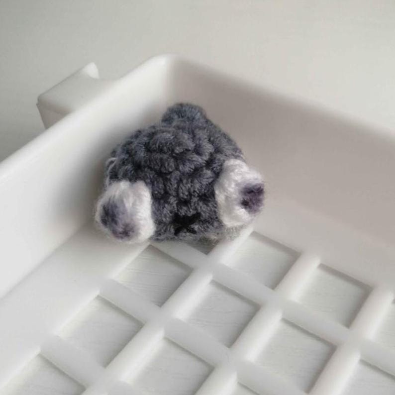 Puede incluir: Un art&iacute;culo tejido a mano, gris y blanco, posiblemente un peque&ntilde;o trasero de animal, con una cruz negra cosida en la parte inferior. El art&iacute;culo se coloca en una bandeja de pl&aacute;stico blanco.