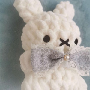 May include: A white crocheted bunny with a gray and white lace bow and a pearl accent. The bunny has black eyes and a stitched X for a mouth.