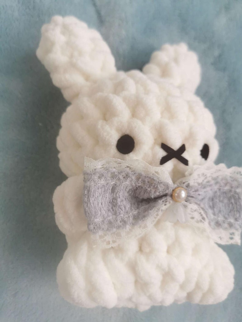 May include: A white crocheted bunny with a gray and white lace bow and a pearl accent. The bunny has black eyes and a stitched X for a mouth.