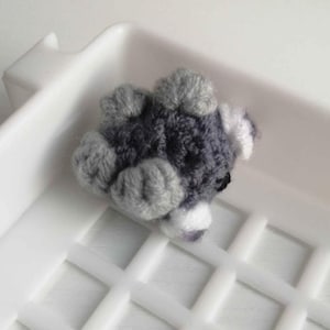 Puede incluir: Peque&ntilde;a figura de animal hecha a mano, de ganchillo, en tonos grises y blancos. El juguete de peluche est&aacute; tumbado, con las patas visibles. El objeto est&aacute; sobre un fondo blanco.
