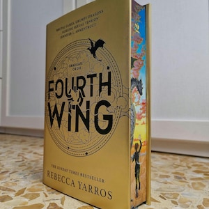 Op de afbeelding: Een hardcover boek getiteld "Fourth Wing" van Rebecca Yarros. Het boek heeft een gouden kaft met zwarte tekst en illustraties, waaronder een draak en de woorden "Graduate or Die". De rand van het boek heeft een kleurrijke illustratie.