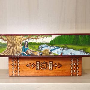 May include: A hand-painted book cover depicting a couple relaxing by a river in a forest setting. The woman is wearing a blue dress and the man is wearing a kilt. The cover is mounted on a wooden box with intricate carvings.