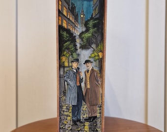 May include: A decorative bookend featuring a nighttime street scene with two men in coats and hats. The background shows buildings, streetlights, and a clock tower under a moonlit sky. The bookend has a wooden frame.