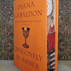 Puede incluir: Un libro de tapa dura naranja con el título "Dragonfly in Amber" de Diana Gabaldon, la autora más vendida del New York Times de "Outlander". La portada del libro presenta un cáliz dorado y una mujer con un vestido rojo.